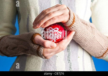 young woman against blue background wearing winter clothes holding red christmas ball toy in her hands Stock Photo