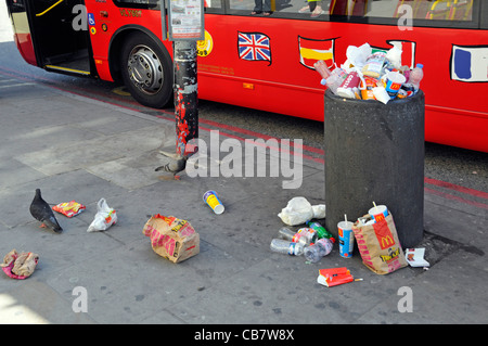 Waste management required to littering scene street refuse bin full of rubbish garbage litter & trash spilling on pavement beside London tour bus UK Stock Photo