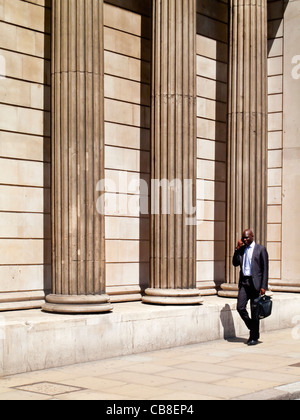 Worker in the City of London walking past the columns of the Bank of England in Threadneedle Street and using mobile phone Stock Photo