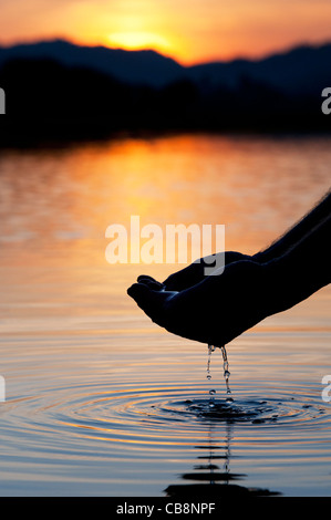 Cupped hands scooping up water in a still lake at sunrise in India. Silhouette Stock Photo