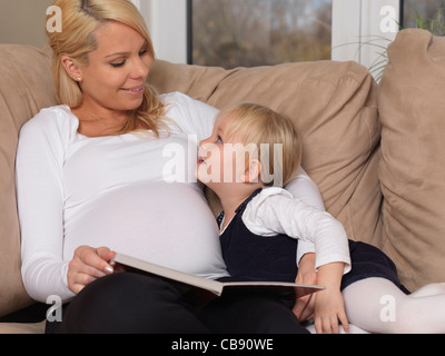 Young pregnant mother and her three year old daughter reading a book together at home Stock Photo