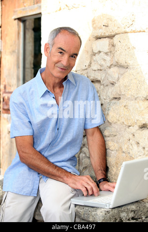 Grey haired man sat by stone wall with laptop Stock Photo