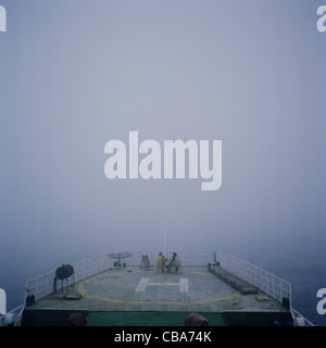 Crew sitting on the helicopter landing pad deck of a ship, in the fog, at sea.