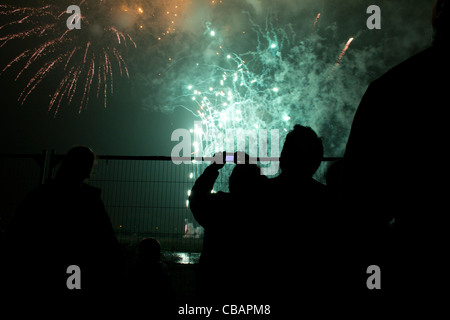 Fireworks display in London, bright green and red explosions fill the sky while silhouetted people look from behind a fence Stock Photo