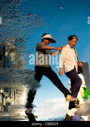 Reflection in a puddle of  a couple trying to avoid wet feet. In the background a skyscraper against a blue sky. Stock Photo