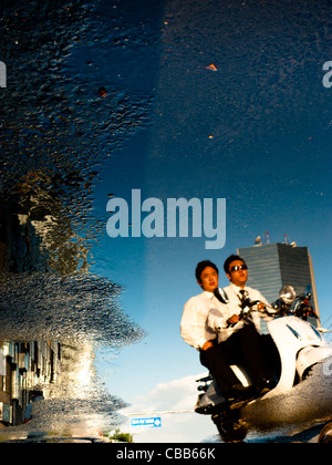 'California Dreaming'. Reflection in a puddle of two men on a scooter with, in the background, a skyscraper against a blue sky. Stock Photo