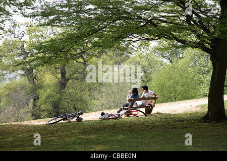 A young couple relax sitting on a hilltop bench in London's Richmond Park. Their bikes rest on the ground beside them. Stock Photo