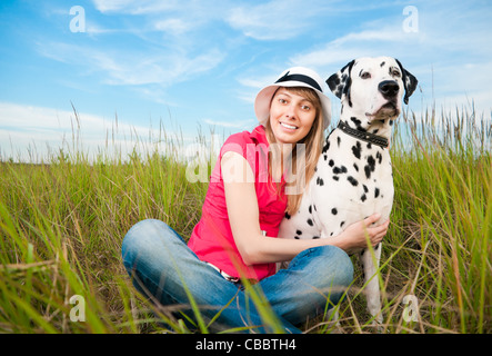 beautiful young woman in hat sitting in grass with her dalmatian dog pet, smiling and looking into the camera Stock Photo