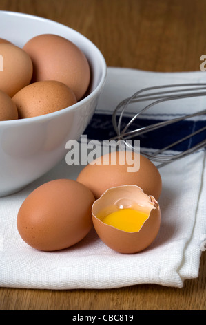 brown hens eggs on a white tea towel with a stainless steel whisk one egg cracked and separated showing egg white and yolk Stock Photo