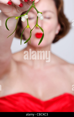 Photo of a woman in a red dress holding some mistletoe and kissing with her eyes shut. Stock Photo