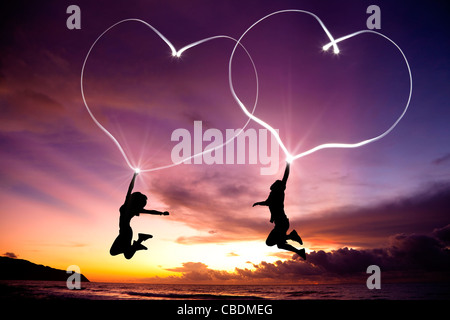 young couple jumping and drawing connected hearts by flashlight in the air on the beach before sunrise Stock Photo