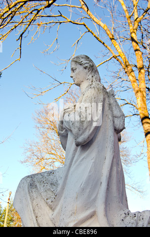 Dirty old white angel statue without wings. Trees without leaves in autumn. Stock Photo
