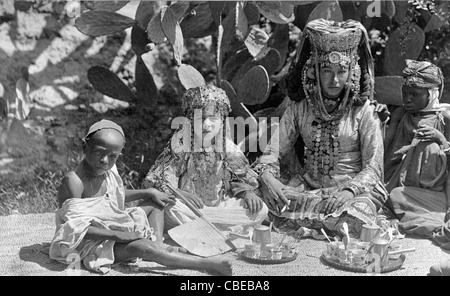 Woman, seated on mat on ground, with young girl and two boys, Algeria. The woman and her daughter are wearing coin necklaces and elaborate headdresses, circa 1890 Stock Photo