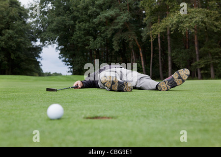 A distraught golfer lying on putting green with ball at the edge of hole Stock Photo