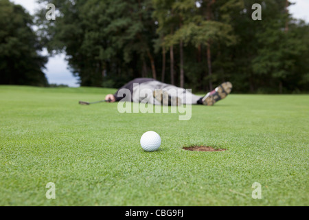 A distraught golfer lying on putting green with ball at the edge of hole Stock Photo