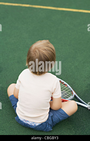 A young boy sitting on a tennis court with a tennis racket, rear view Stock Photo