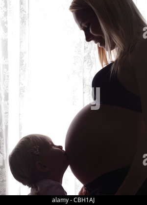 Little daughter kissing her happy pregnant mother's belly in bright light in front of a window