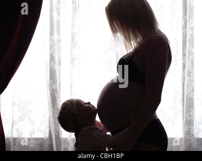 Happy little daughter hugging her pregnant mother's belly in bright light in front of a window Stock Photo