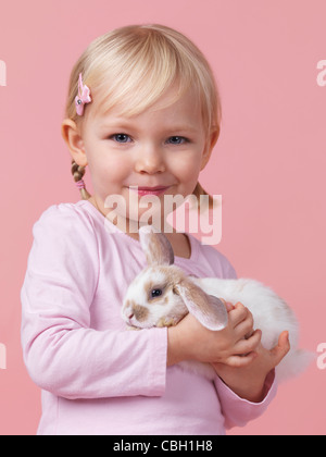 Portrait of a three year old smiling girl holding a pet rabbit in her hands isolated on pink background Stock Photo