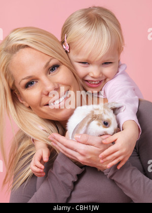 Portrait of a smiling three year old girl with her mother holding a pet rabbit in her hands isolated on pink background Stock Photo