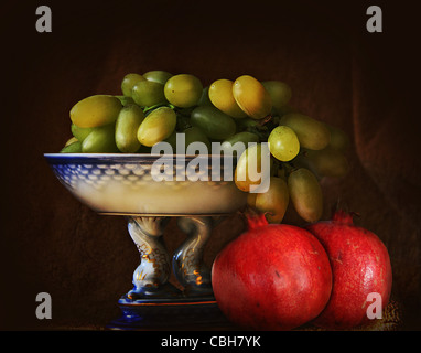 Retro still life, porcelain tray with grapes and two pomegranates in chiaroscuro on a dark grunge background Stock Photo