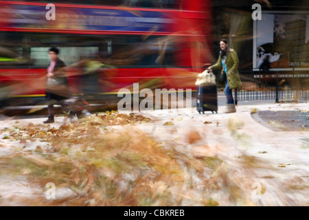 people caught in a gust of wind with leaves swirling around and a bus passing Stock Photo