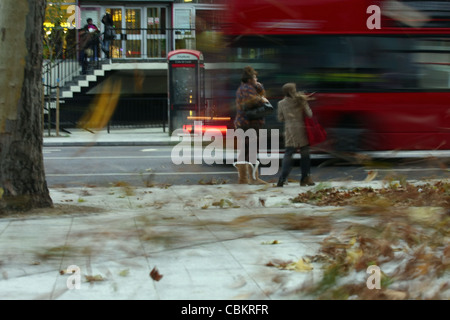 Two females caught in a gust of wind with leaves swirling around and a bus passing Stock Photo