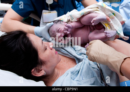 Mother in labor giving birth to baby new life in hospital. Infant covered in grease and lanugo is crying and held by nurses. Stock Photo