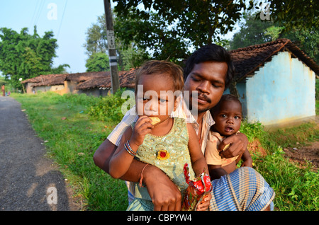 Indian adult man holding two young children and sitting on ground in a sunny morning near a hut in the countryside. Chhatisgarh, India Stock Photo