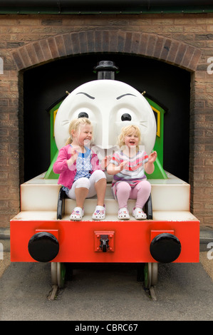 Vertical portrait of two little girls laughing and having fun sitting on a train character at an amusement park in the sunshine. Stock Photo