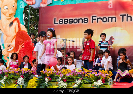 A little girl and boy sing into mikes while young children watch them on stage during school performance in Chiang Mai Thailand Stock Photo