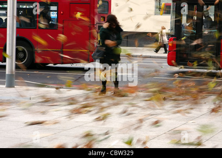 Leaves swirling around a female caught in a sudden gust of strong wind, with red London buses passing in the background Stock Photo