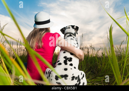 beautiful young woman in hat sitting in grass with her dalmatian dog pet with their backs to camera. Sunset cloudy sky Stock Photo