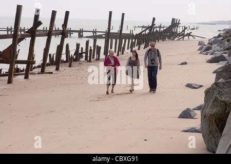 Happisburgh coastline, North Norfolk, East Anglia. Three people walking between failing timber and metal breakers, groynes; imported rocks right Stock Photo