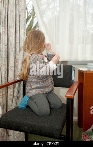 female toddler sat on chair looking out of window holding curtain