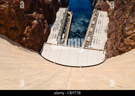 Looking down from the top of the massive concrete wall of the Hoover Dam. At top is the outflow of the Colorado River with the twin buildings housing the hydroelectric turbines on either side of the river. The dam spans the state border, with Arizona on the left and Nevada on the right. Stock Photo