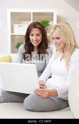 Two beautiful women at home sitting on sofa or settee using laptop computer and smiling Stock Photo