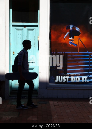 A silhouetted youth, with skateboard under arm, passes some advertising with a skateboarder in full action on Stock Photo