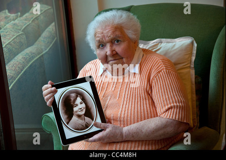 98 years old elderly lady holding an iPad tablet computer, displaying a sepia portrait of herself taken 80 years ago at age 18 Stock Photo