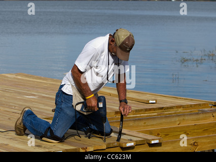 A carpenter fastening a board down on a dock out over a lake using an electric drill. Stock Photo