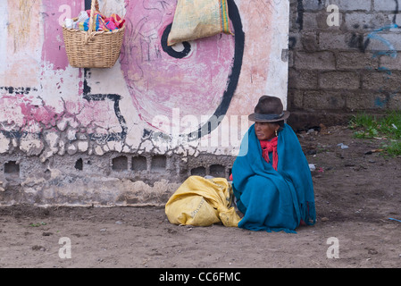A very old female Andean Indian sits on the ground near a graffiti marked wall in Saquisili, Ecuador, South America. Stock Photo