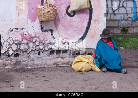 A very old female Andean Indian sits on the ground near a graffiti marked wall in Saquisili, Ecuador, South America. Stock Photo