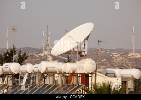 Solar heating panels and tanks, large satellite dish on roof with wind turbines behind. Pervolia, Cyprus Stock Photo