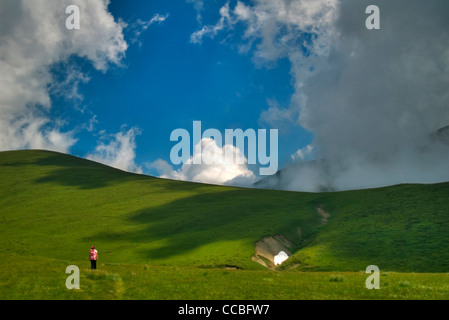 Female hiker walking on lush green high mountain meadows under overhanging white fluffy clouds Stock Photo