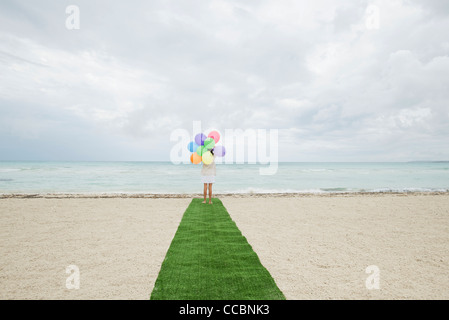 Girl standing on artificial turf on beach with bunch of balloons, rear view