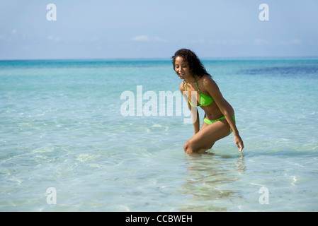 Young woman in bikini standing in ocean, bending to touch water, portrait Stock Photo