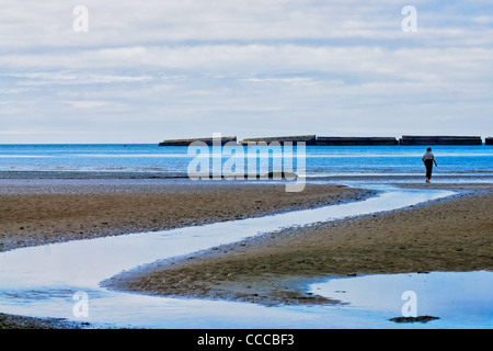 Arromanche, Normandy, France. A man walks on the seashort. Remnants of the 'Mulberry' Artificial harbor from WWII in the back. Stock Photo