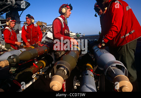 Red shirted ordnance men prepare to fit smart bombs and missiles to an F/A-18 fighter jet on deck of USS Harry S Truman. Stock Photo