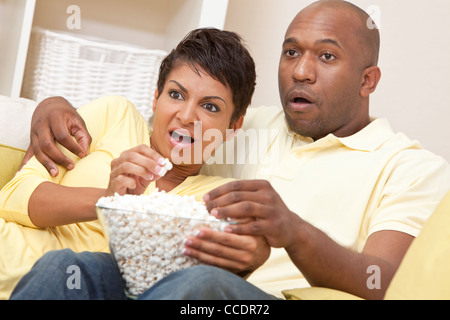 A happy African American man and woman couple in their thirties sitting at home, eating popcorn and looking shocked Stock Photo