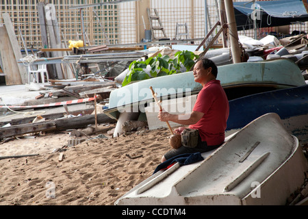 Man sitting on an upturned fishing boat playing a sitar on St Stephens Beach, Stanley Hong Kong SAR China Stock Photo